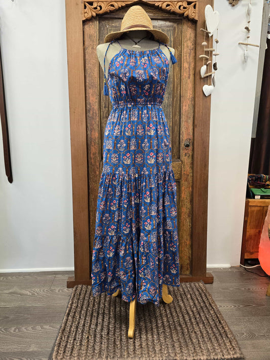 A sleeveless tiered maxi dress with a blue base and red, white, and yellow floral pattern, displayed on a mannequin with a straw hat in front of a rustic wooden door.