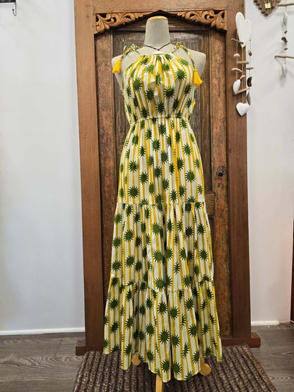 A sleeveless tiered maxi dress with a yellow and white vertical stripe pattern and green palm tree print, displayed on a mannequin in front of a rustic wooden door.