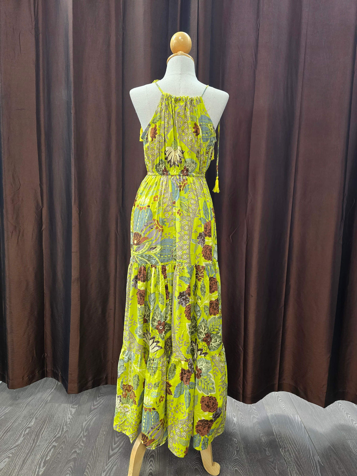 A sleeveless, tiered maxi dress with a vibrant yellow-green base and a multicolor floral and paisley print, displayed on a mannequin in front of a dark brown curtain backdrop.