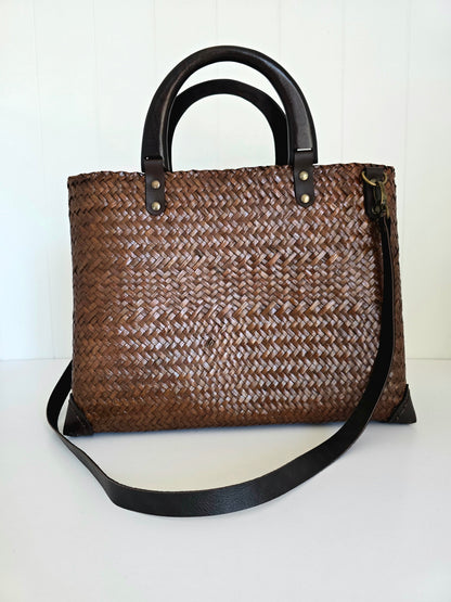 Brown woven handbag with black handles and crossbody strap on a white background
