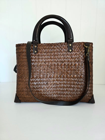 A brown woven tote bag with dark brown wooden handles, trim, and a detachable faux leather shoulder strap, shown against a white paneled background.
