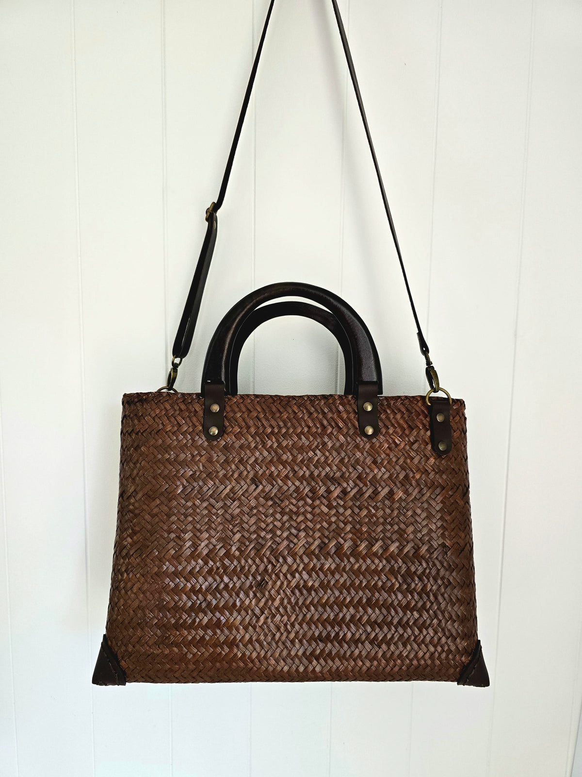 A brown woven straw tote bag with black leather handles and a long black shoulder strap, hanging against a white paneled wall.