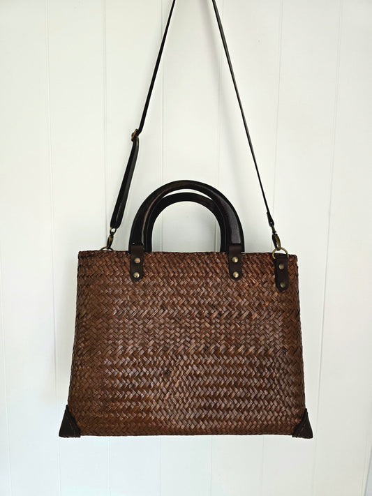 A brown woven straw tote bag with black leather handles and a long black shoulder strap, hanging against a white paneled wall.