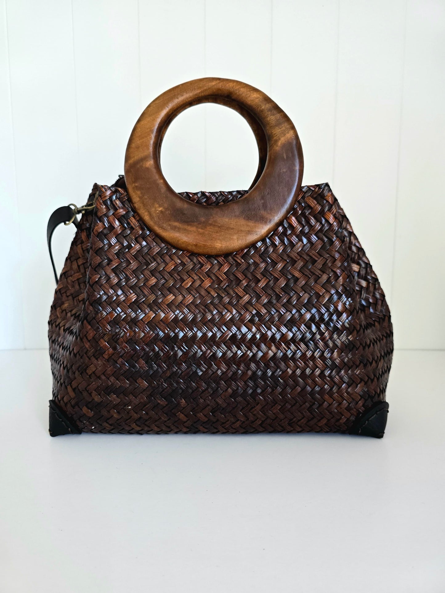 A dark brown woven Natural fibre handbag with a large circular wooden top handle, set against a white paneled background.