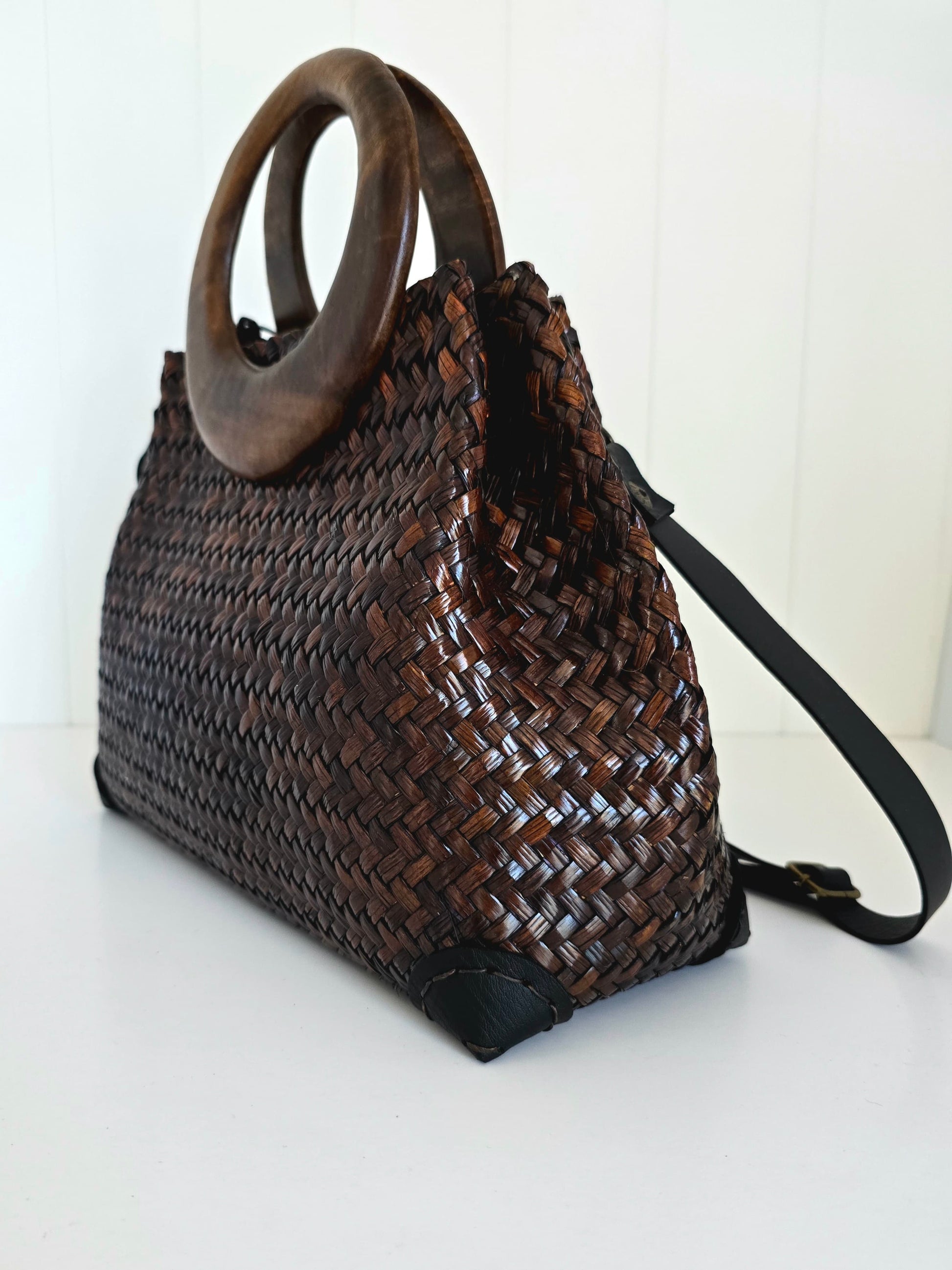 A dark brown woven natural fibre handbag with a large circular wooden top handle and a black shoulder strap, shown on a white surface against a white paneled background.