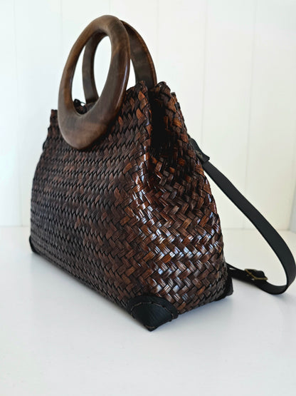A dark brown woven natural fibre handbag with a large circular wooden top handle and a black shoulder strap, shown on a white surface against a white paneled background.