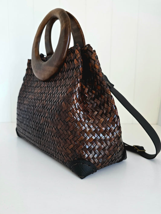 A dark brown woven natural fibre handbag with a large circular wooden top handle and a black shoulder strap, shown on a white surface against a white paneled background.