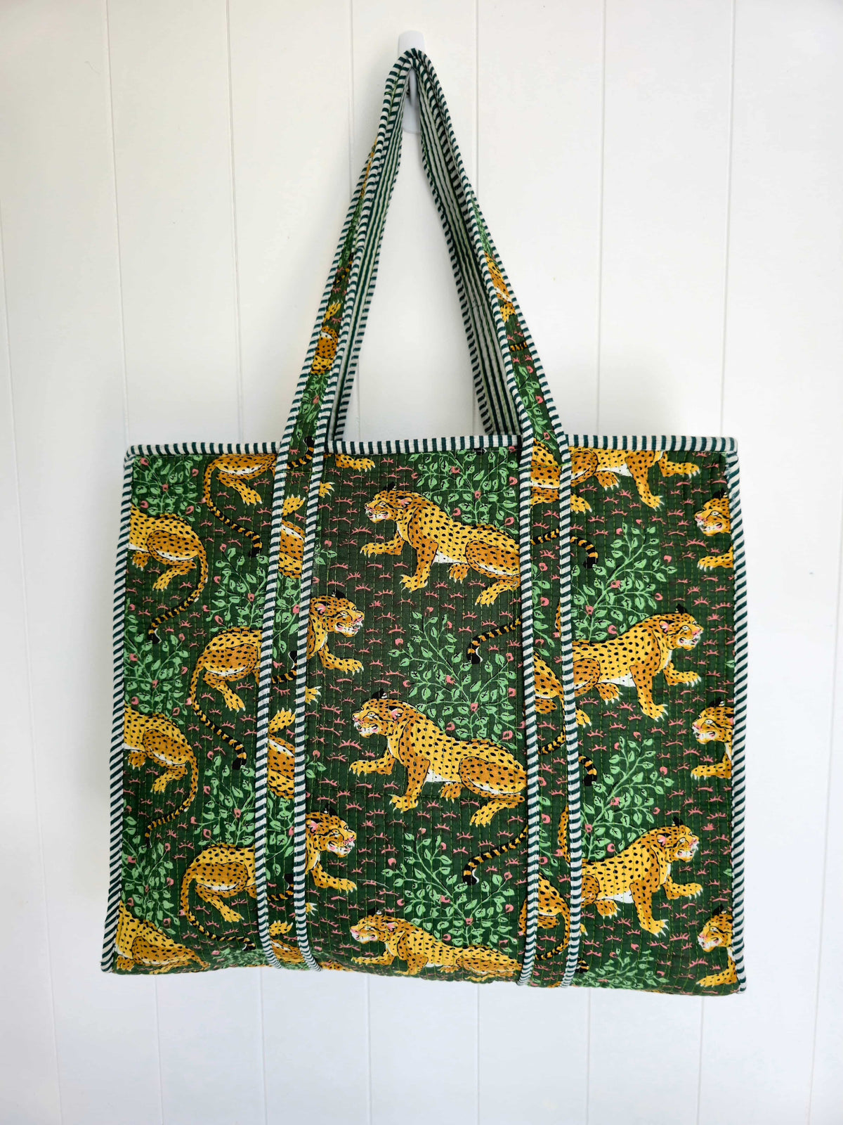 A large tote bag featuring a green and black jungle print with yellow leopards, hanging against a white paneled wall.