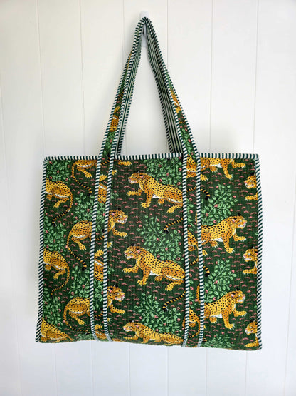 A large tote bag featuring a green and black jungle print with yellow leopards, hanging against a white paneled wall.