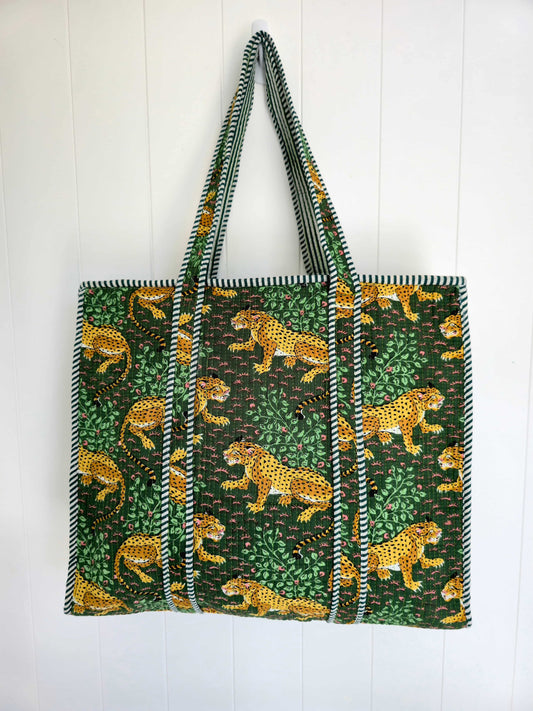 A large tote bag featuring a green and black jungle print with yellow leopards, hanging against a white paneled wall.