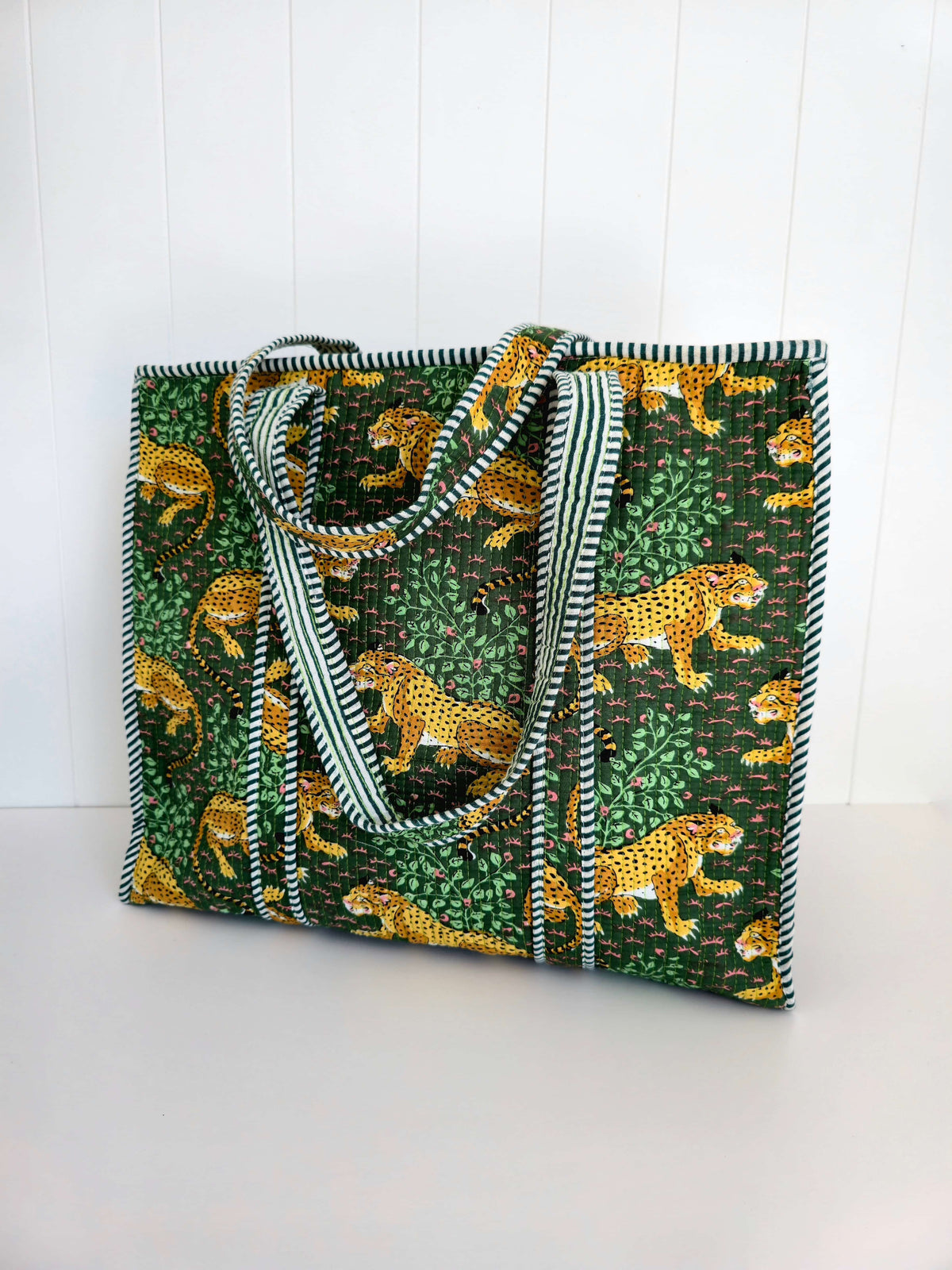 A large tote bag featuring a green and yellow animal print with cheetahs and leafy patterns, complete with striped green and shoulder straps, set against a white paneled background.