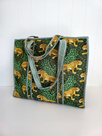 A large tote bag featuring a green and yellow animal print with cheetahs and leafy patterns, complete with striped green and shoulder straps, set against a white paneled background.