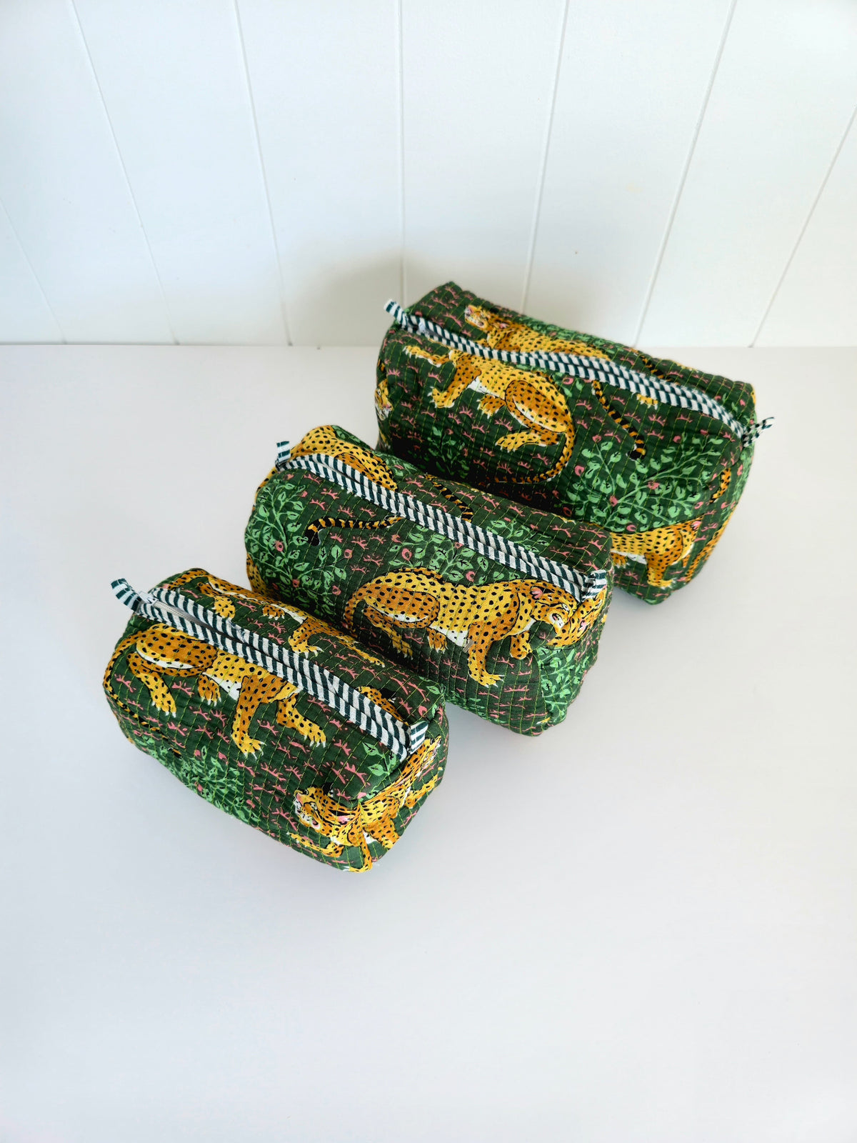 Three zippered cosmetic pouches in green and gold leopard-print fabric, arranged diagonally on a white surface against a white paneled background.