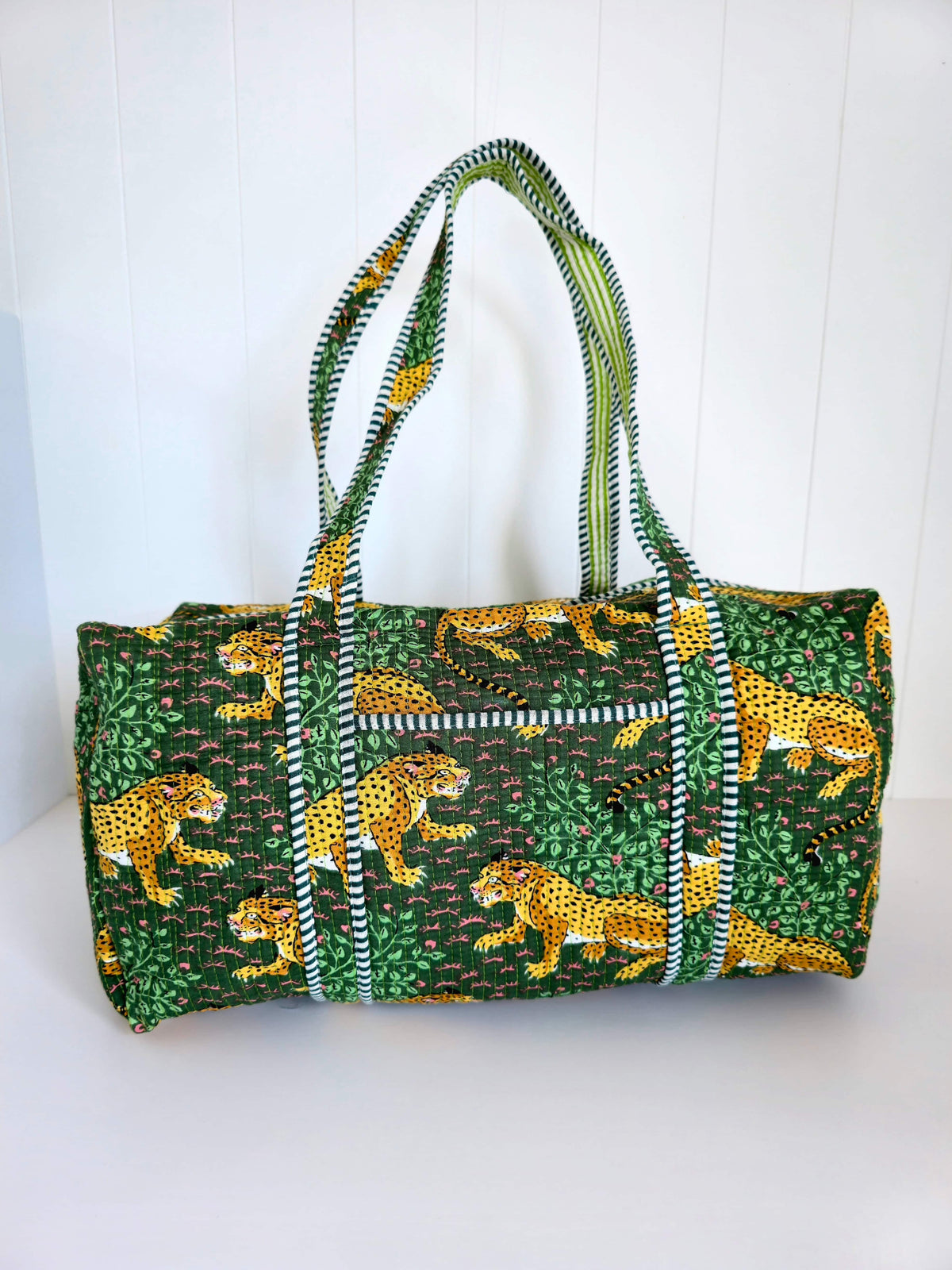 A cylindrical weekender bag with a green and yellow animal print featuring cheetahs and leafy patterns, accented with striped green and white handles and trim, set against a white paneled background.