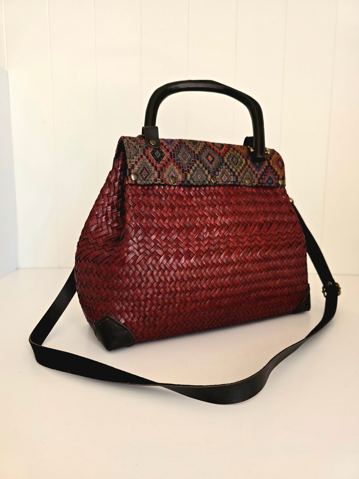 A woven red handbag with a black top handle and shoulder strap, featuring a patterned flap in blue, red, and brown tones.
