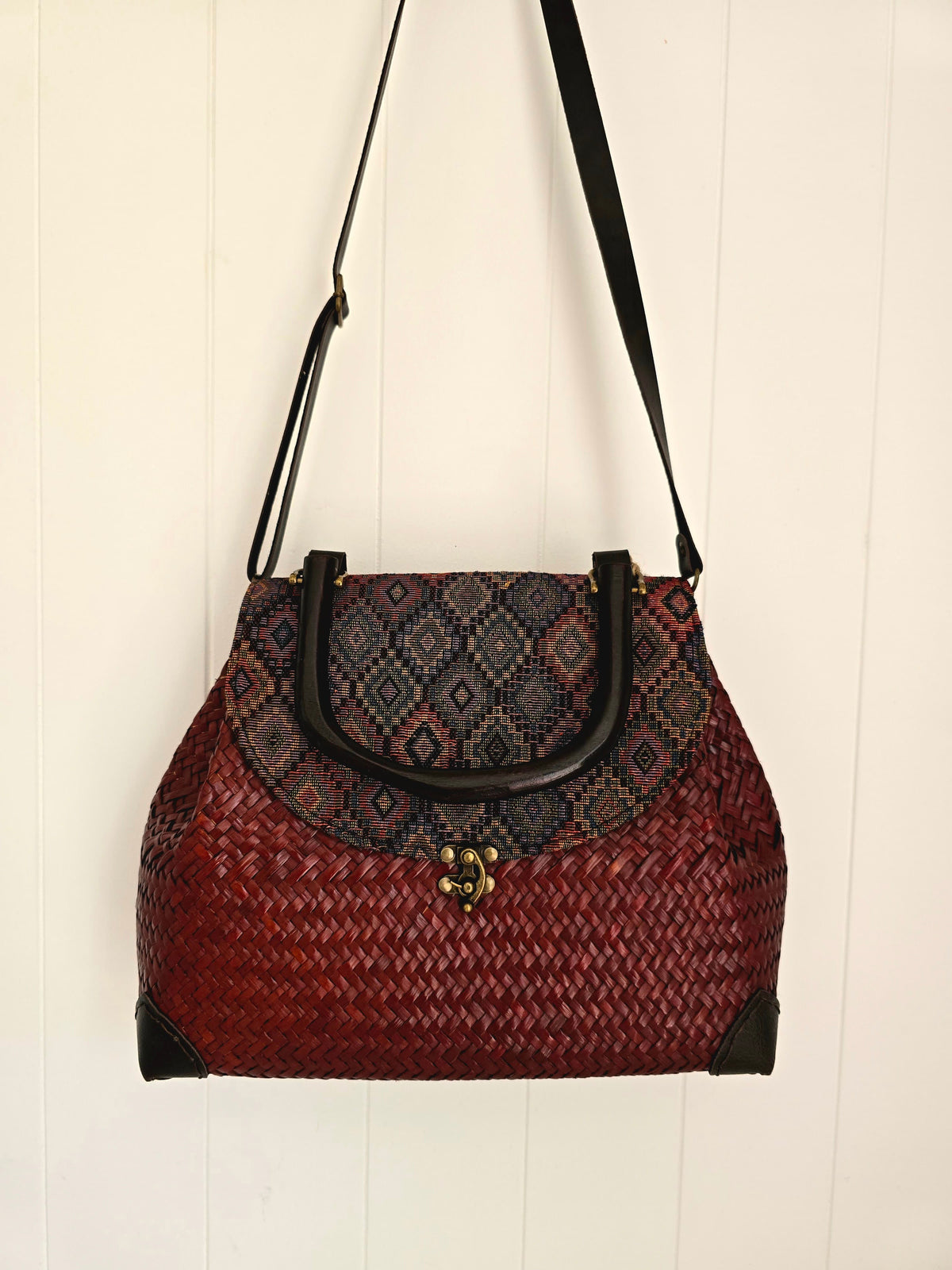 A woven shoulder bag with a red and black patterned body, black trim, and a black shoulder strap, hanging against a white paneled wall.