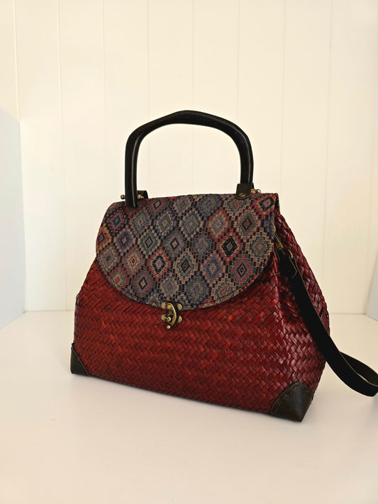 A woven red handbag with a patterned flap in blue, purple, and red, black wooden handles, and a black strap, set against a white paneled background.