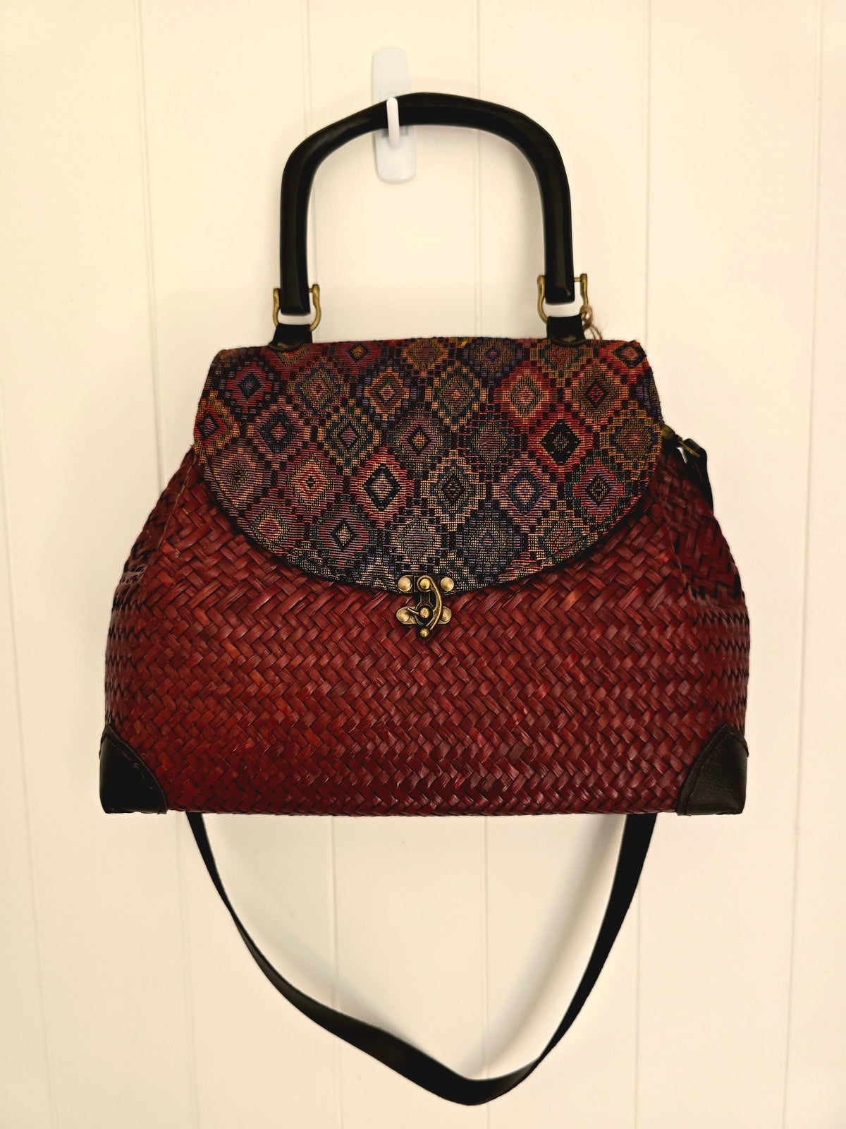 A woven handbag with a dark red body, a patterned flap in shades of red, blue, and brown, black handles and strap, and gold-tone hardware, hanging against a white paneled wall.