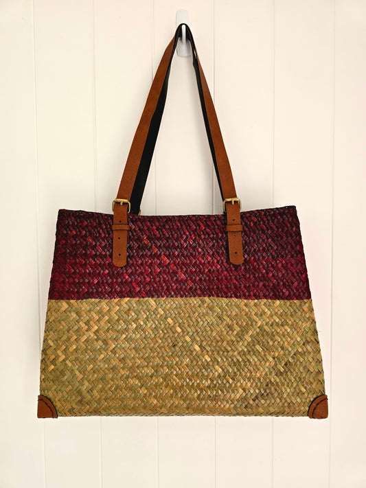 A woven tote bag with a three-tone design, featuring a dark red upper third, lighter red middle third and a natural straw-colored lower half, with brown faux leather shoulder straps and corner protectors, hanging against a white paneled wall.