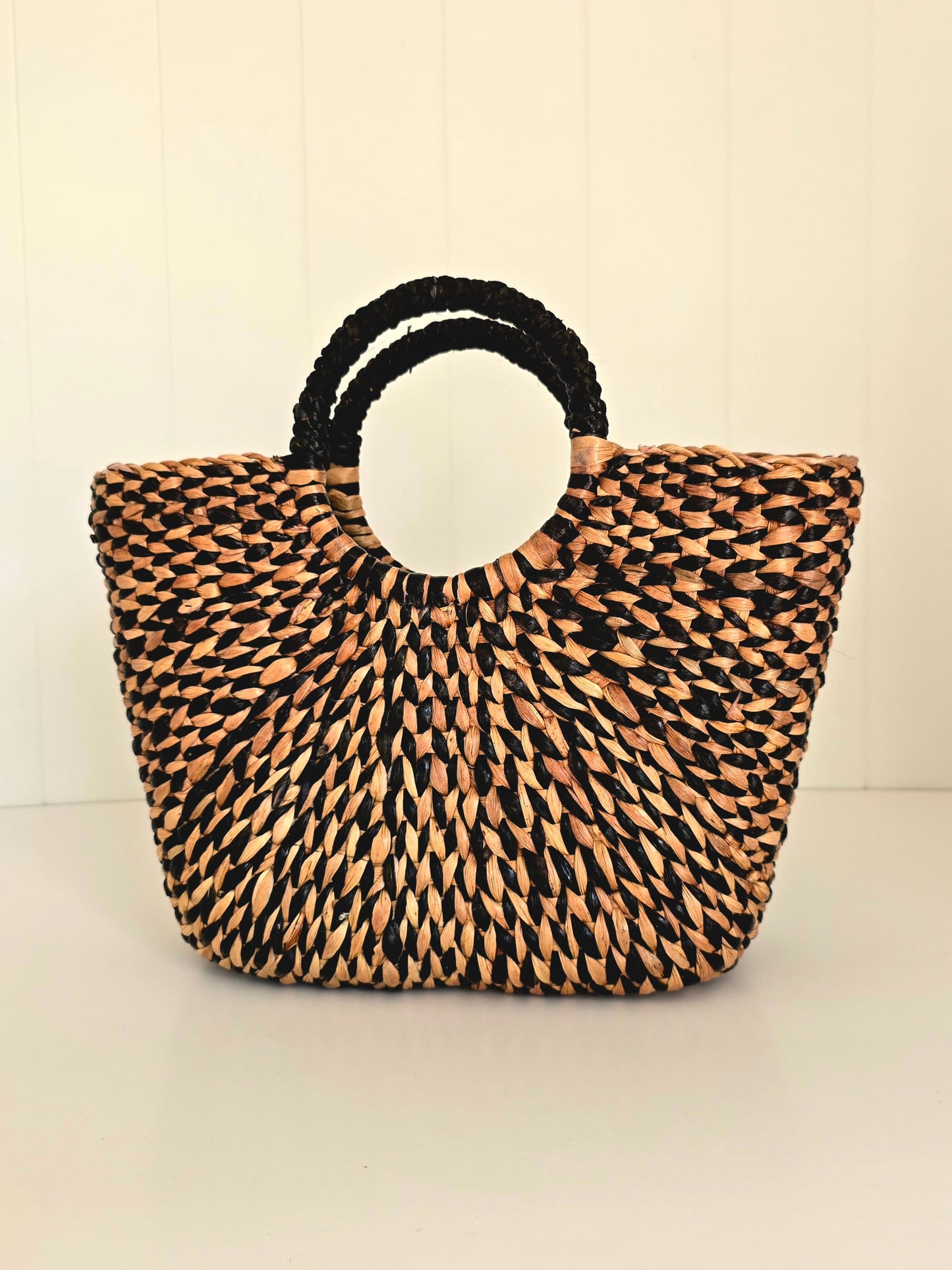 A woven straw tote bag with black braided handles, set against a light cream paneled background.
