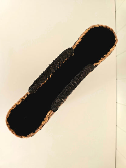 A woven straw basket with a black fabric lining and black braided straps, shown from a top-down perspective on a light beige background.