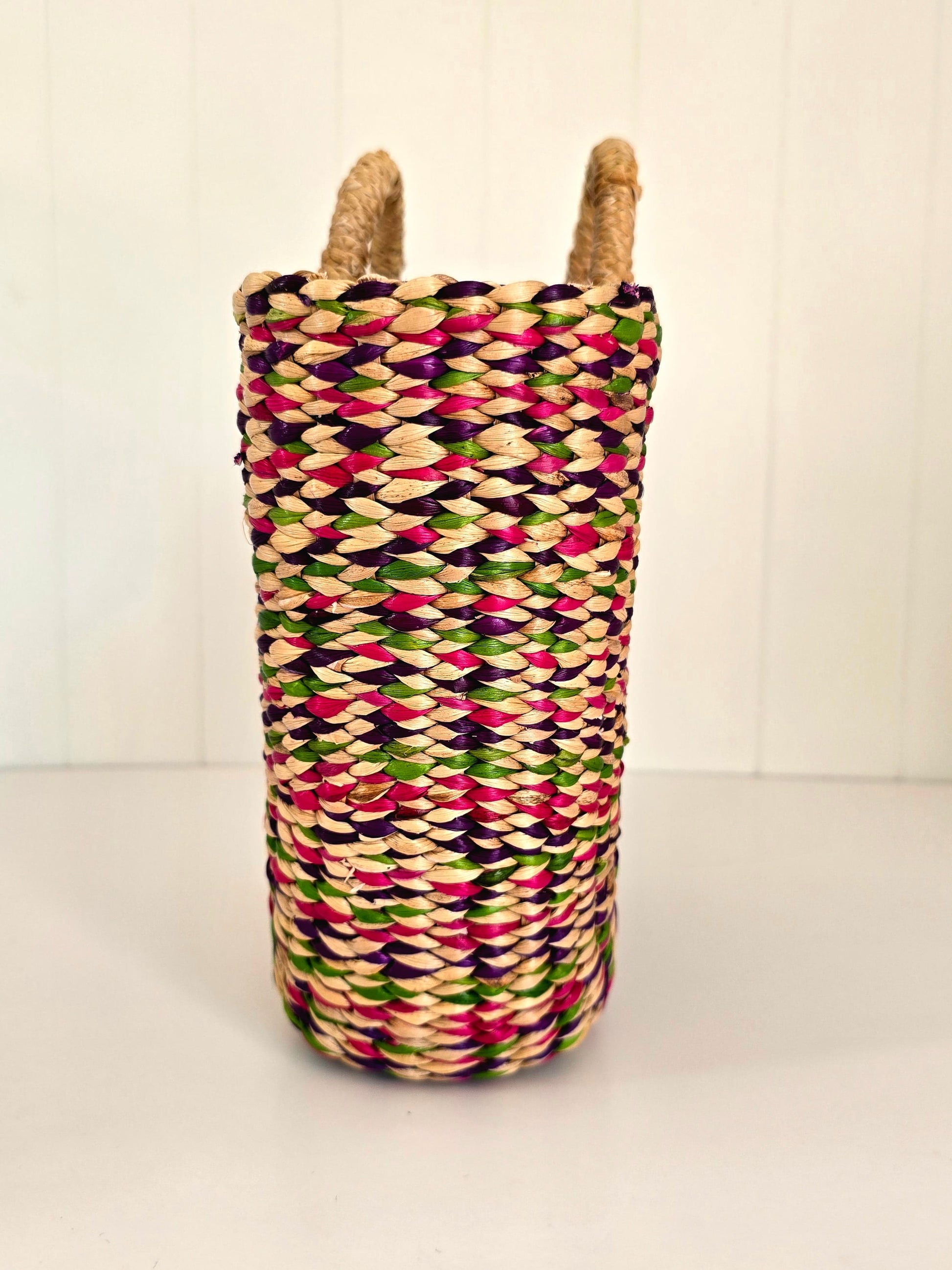 A tall woven basket with two natural jute handles, featuring a colorful pattern of pink, green, purple, and beige woven fibers, set against a white paneled background.