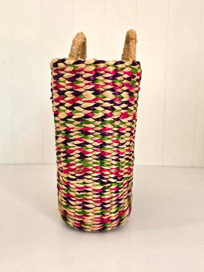 A tall woven basket with two natural jute handles, featuring a colorful pattern of pink, green, purple, and beige woven fibers, set against a white paneled background.