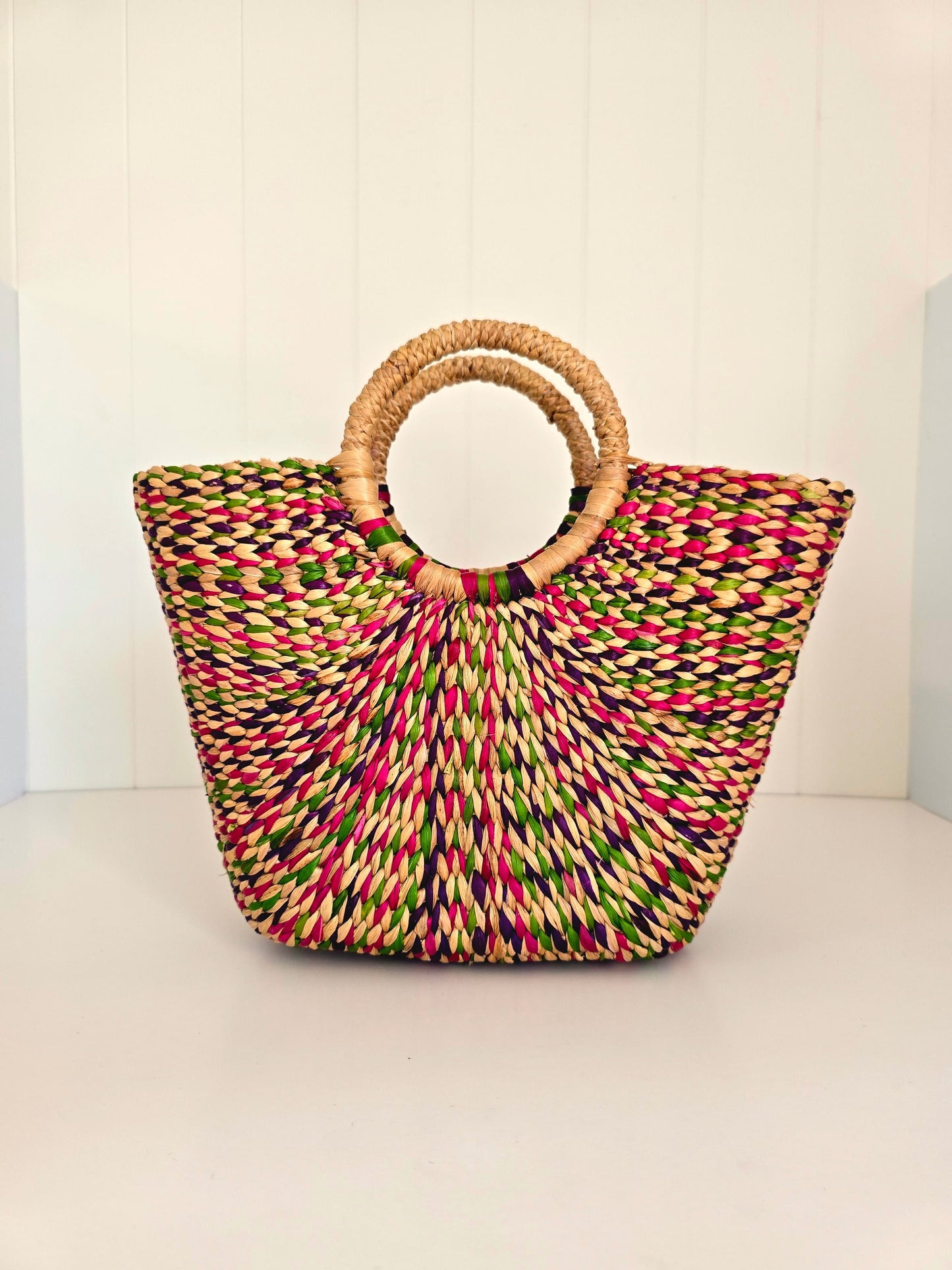 A woven straw tote bag with a multicolor pattern of pink, green, yellow, and black threads, featuring two natural light brown woven handles, set against a white paneled background.