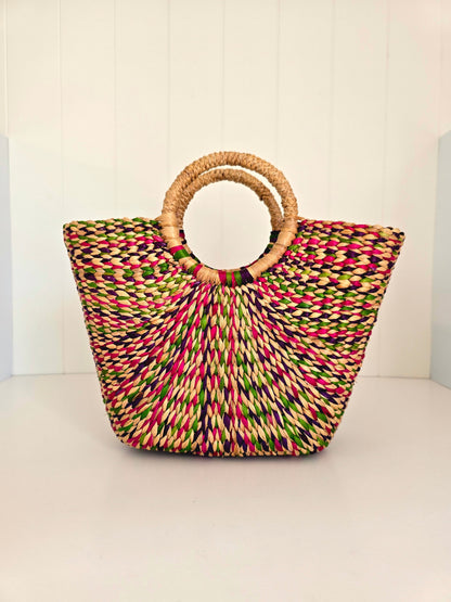 A woven straw tote bag with a multicolor pattern of pink, green, yellow, and black threads, featuring two natural light brown woven handles, set against a white paneled background.