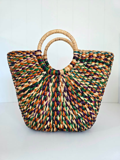 A woven straw tote bag with a circular ring handle, featuring a multicolor pattern of natural beige, green, orange, and purple threads, set against a plain white background.