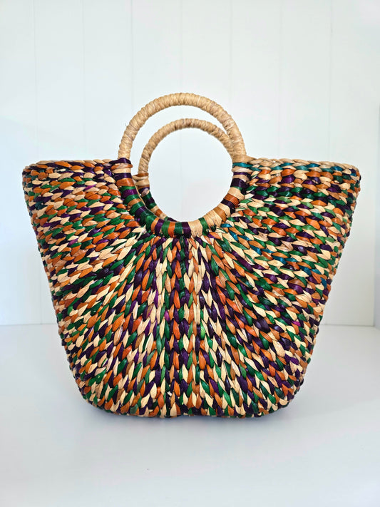 A woven straw tote bag with a circular ring handle, featuring a multicolor pattern of natural beige, green, orange, and purple threads, set against a plain white background.