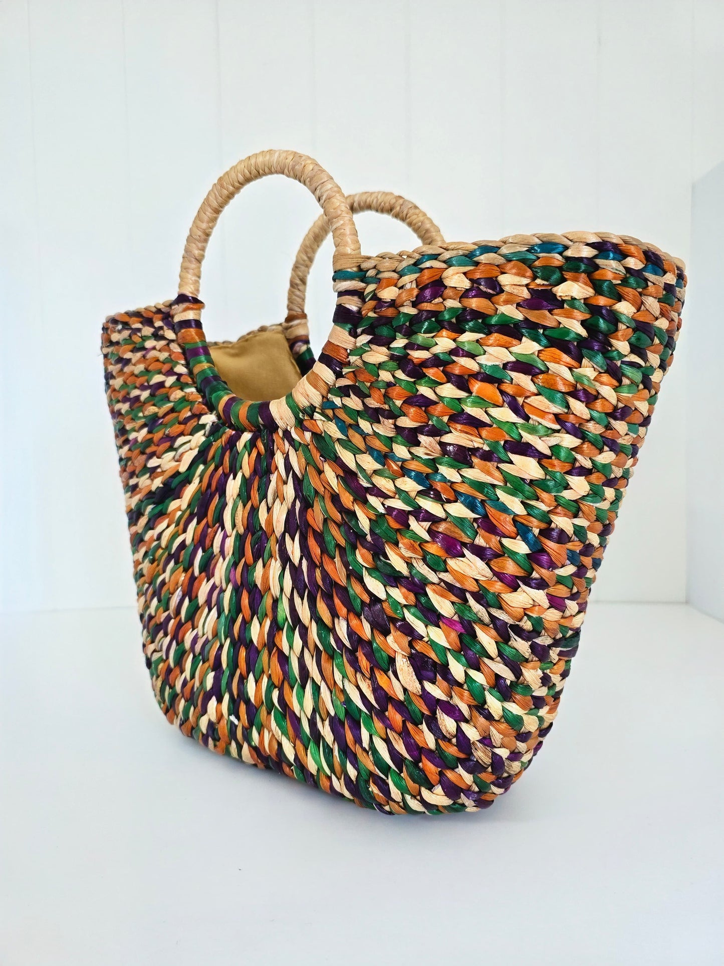 A woven straw tote bag with two round natural-fiber handles, featuring a multicolor pattern of green, orange, purple, and beige woven into a textured body, set against a plain white background.