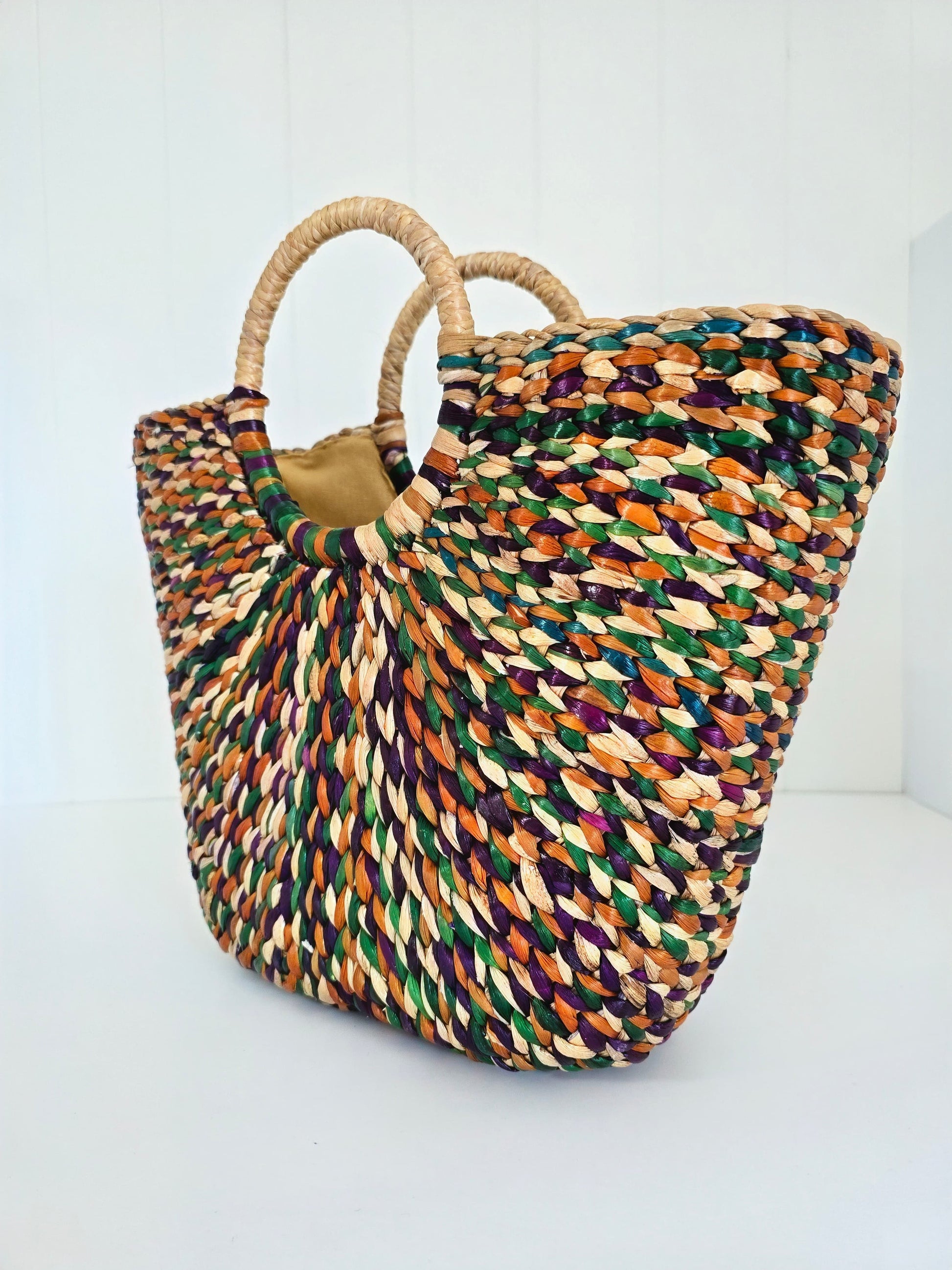 A woven straw tote bag with two round natural-fiber handles, featuring a multicolor pattern of green, orange, purple, and beige woven into a textured body, set against a plain white background.