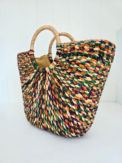 A woven straw tote bag with two round natural-fiber handles, featuring a multicolor pattern of green, orange, purple, and beige woven into a textured body, set against a plain white background.