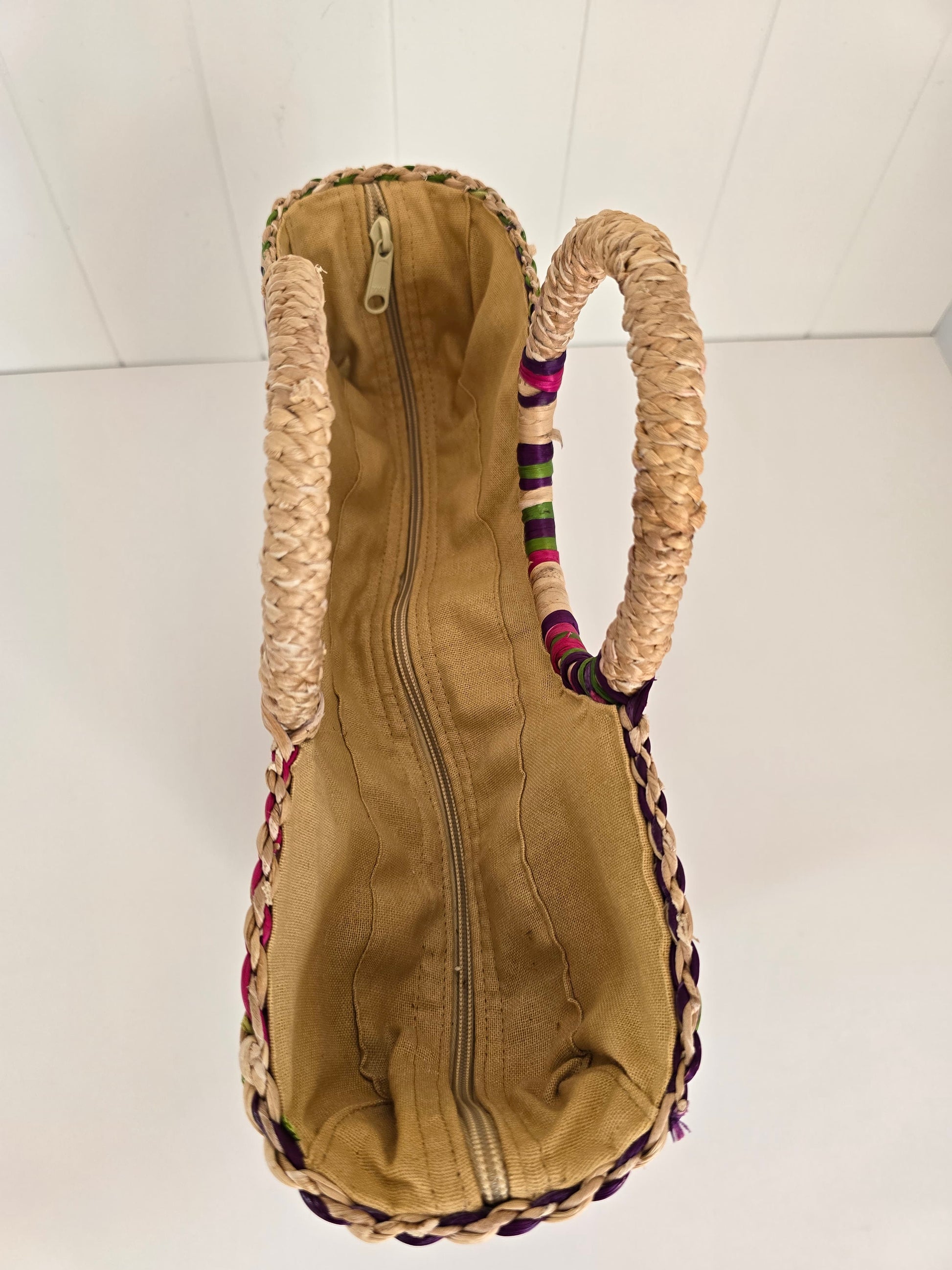 A top-down view of a tan canvas zippered bag with thick braided straw handles and colorful woven trim along the edges, set against a white paneled background.