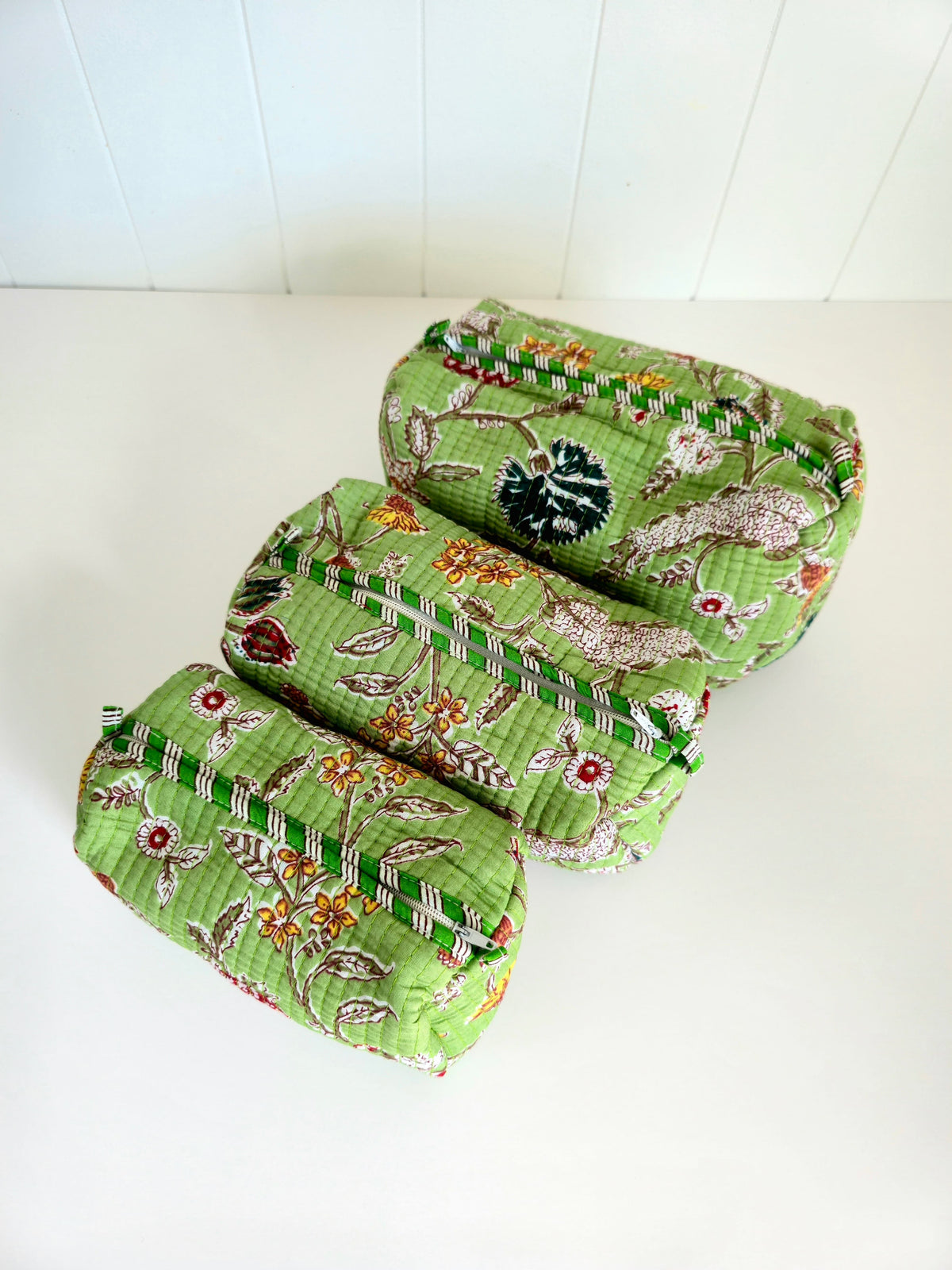 Three small quilted fabric pouches or organizers stacked diagonally on a white surface against a white paneled background. The fabric has a light green base with a floral botanical print in beige, orange, red, and green.