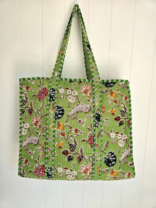 A green woven tote bag with a detailed multicolor floral and bird pattern, hanging against a white paneled wall.