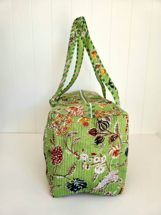 A green quilted fabric weekender bag with a floral pattern, featuring dual shoulder straps, set against a white paneled background.