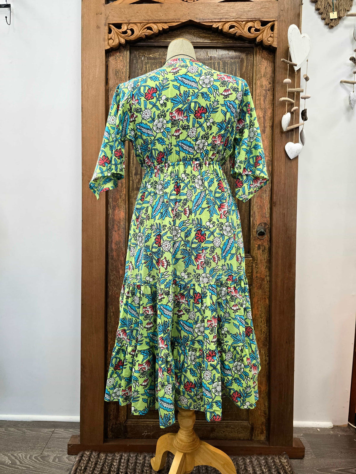 A green tiered midi dress with a colorful floral and leaf print, displayed on a wooden dress form in front of a rustic wooden door.