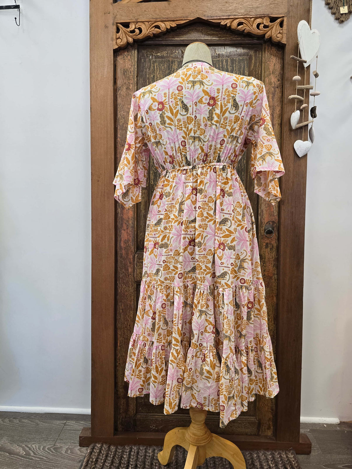A pink and orange floral tiered midi dress displayed on a mannequin from the back, set against a rustic carved wooden door.