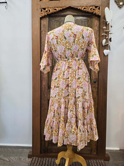 A pink and orange floral tiered midi dress displayed on a mannequin from the back, set against a rustic carved wooden door.