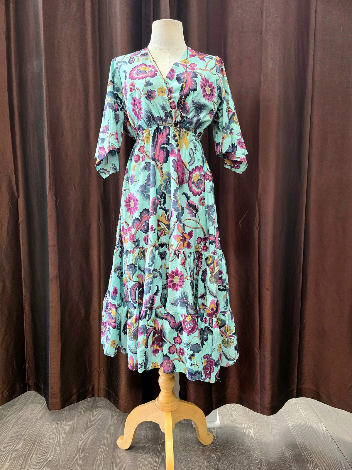 A long-sleeve midi dress with a colorful floral and butterfly print on a mint green base, displayed on a wooden dress form against a brown curtain backdrop.
