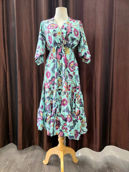A long-sleeve midi dress with a colorful floral and butterfly print on a mint green base, displayed on a wooden dress form against a brown curtain backdrop.