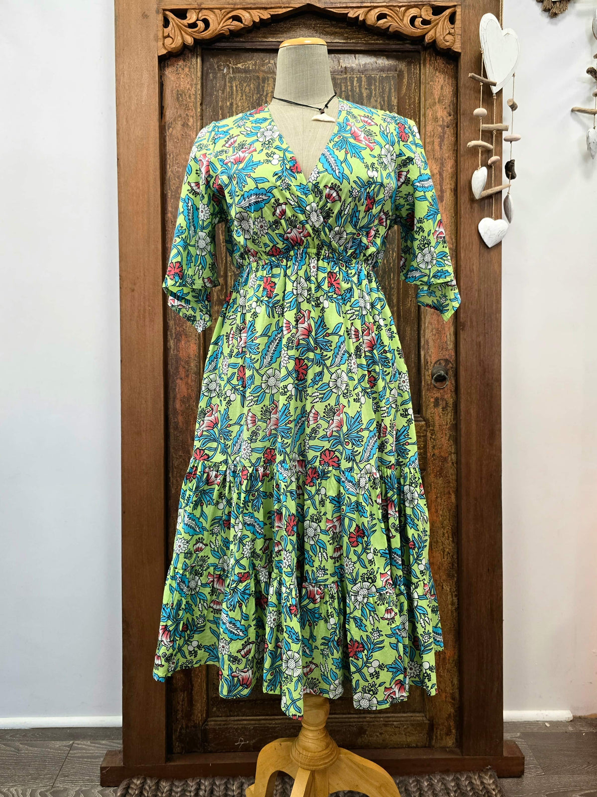 A vibrant green tiered maxi dress with a colorful floral and paisley print, displayed on a mannequin in front of a carved wooden door.