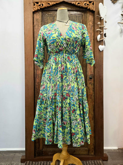 A vibrant green tiered maxi dress with a colorful floral and paisley print, displayed on a mannequin in front of a carved wooden door.