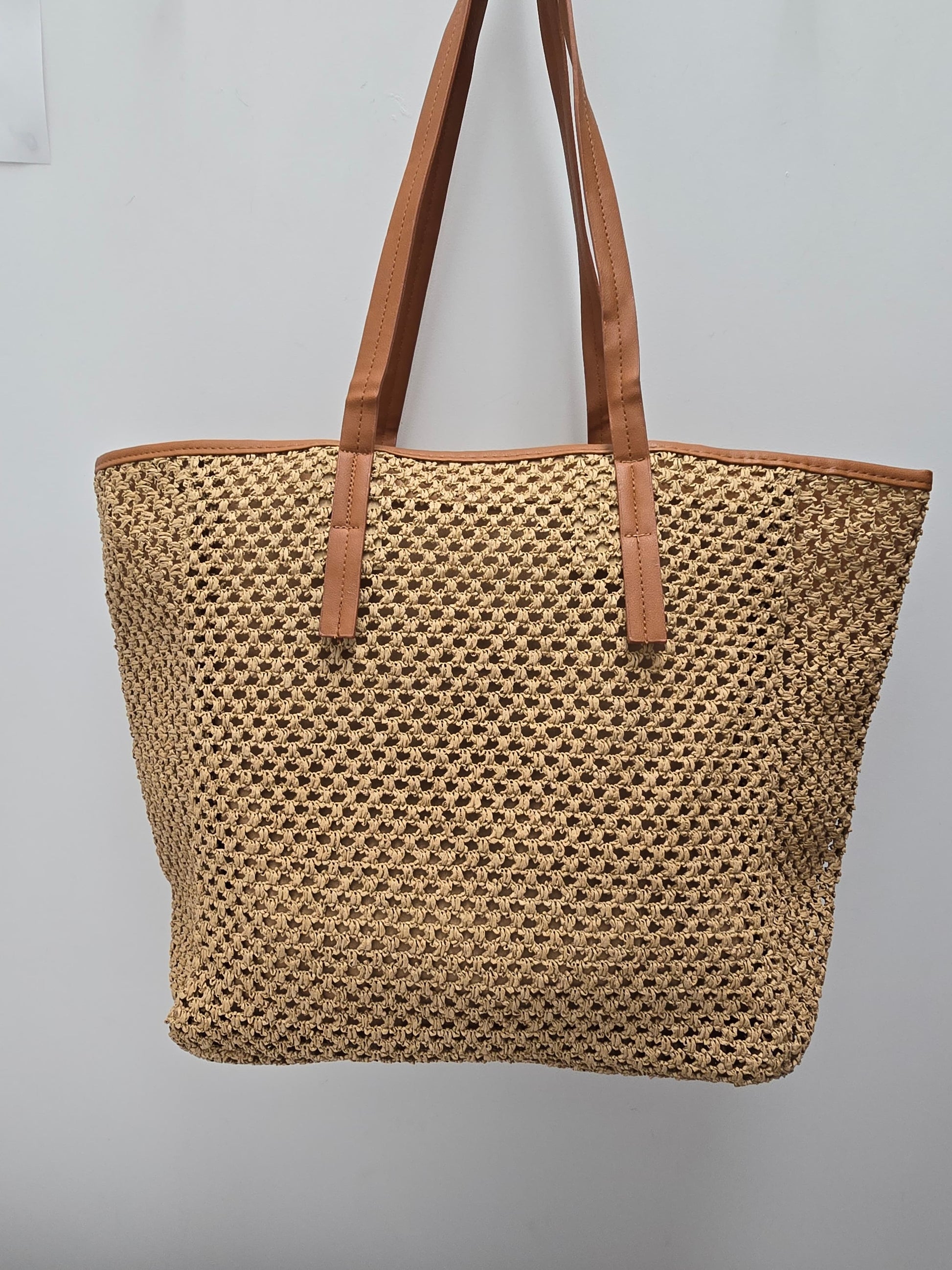 A woven straw tote bag with tan leather shoulder straps and trim, hanging against a plain light gray wall.
