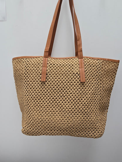 A woven straw tote bag with tan leather shoulder straps and trim, hanging against a plain light gray wall.
