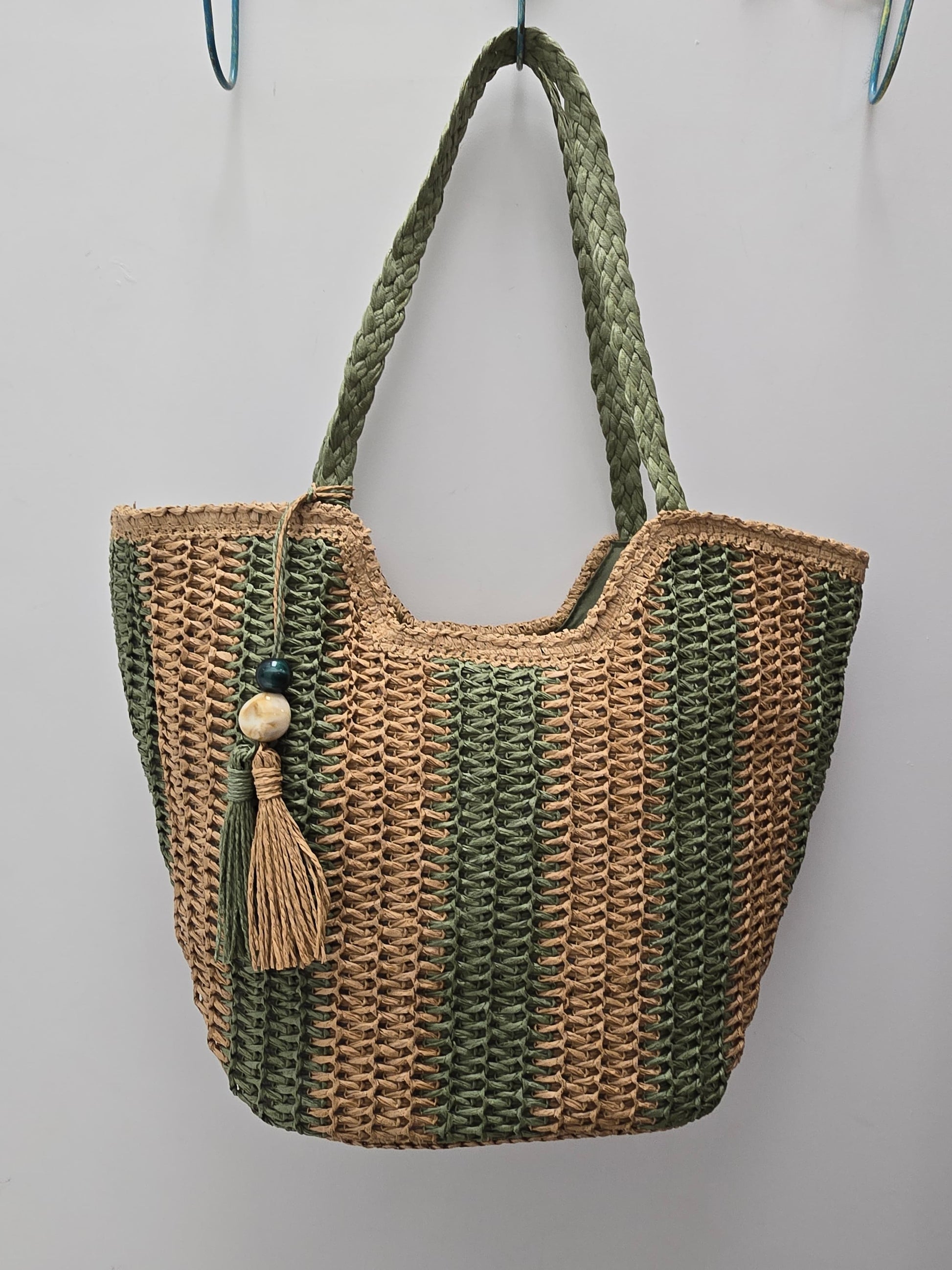 A woven straw and olive green tote bag with braided shoulder straps and a hanging tassel ornament, shown against a plain light gray wall.