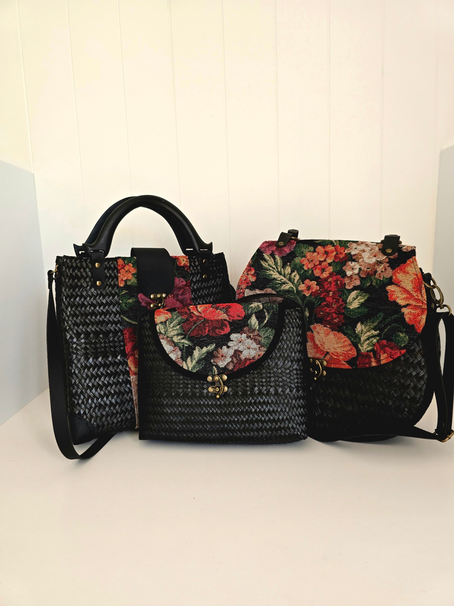 Three black handbags with floral tapestry patterns on black woven body's with faux leather straps and wooden handles, set against a white background
