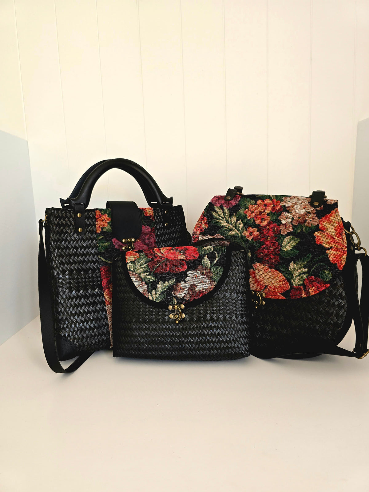 Three black handbags with floral tapestry patterns on black woven body's with faux leather straps and wooden handles, set against a white background